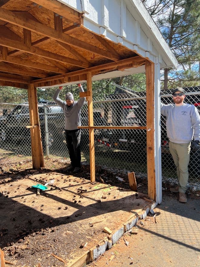 Crew members taking down the frame of a small carport, dump trailer behind the fence