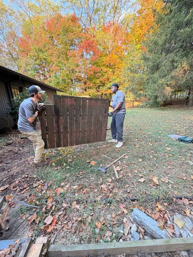 Two crew members carrying an old stained fence panel across a backyard in fall