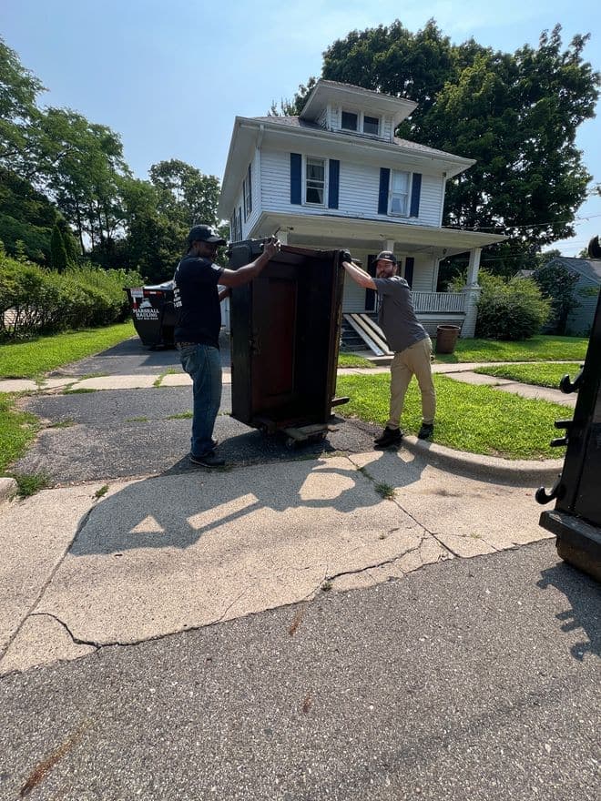 Two crew members wheeling an upright piano down a residential driveway on a dolly