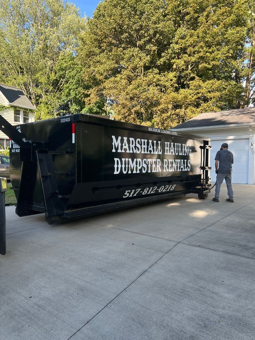 A lettered Marshall Hauling Dumpster Rentals can set on a residential concrete driveway, crew member alongside