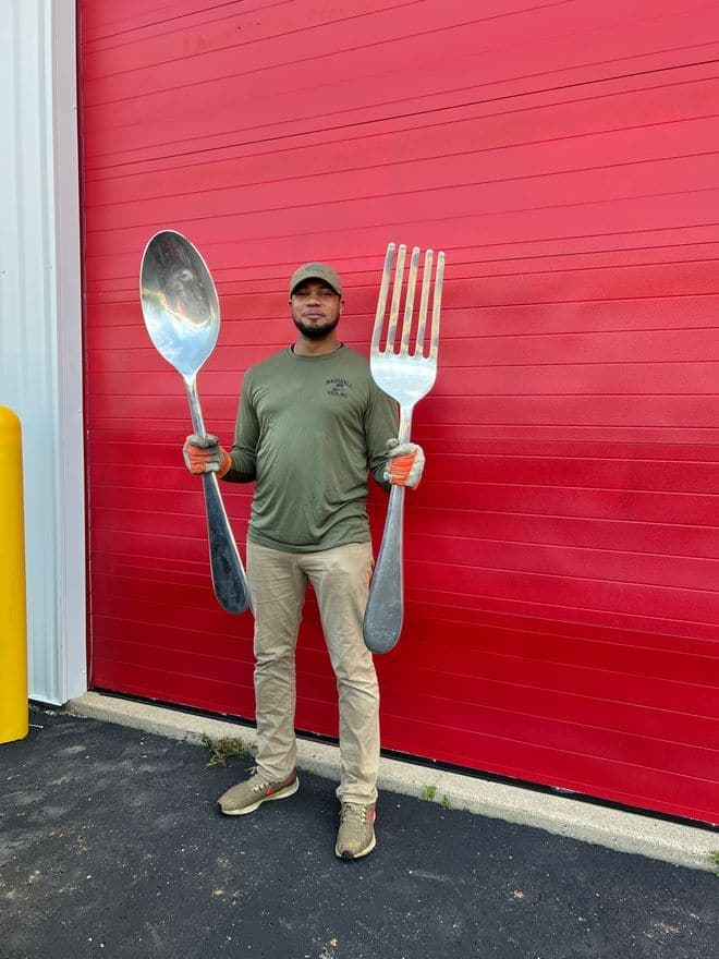A crew member holding a giant novelty spoon and fork in front of a red garage door